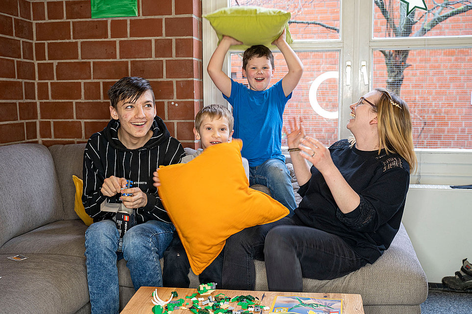 Vier Personen auf dem Sofa: drei Kinder/Jugendliche und eine Frau spielen mit Kissen, lachen und bauen mit Spielzeug auf dem Tisch.
